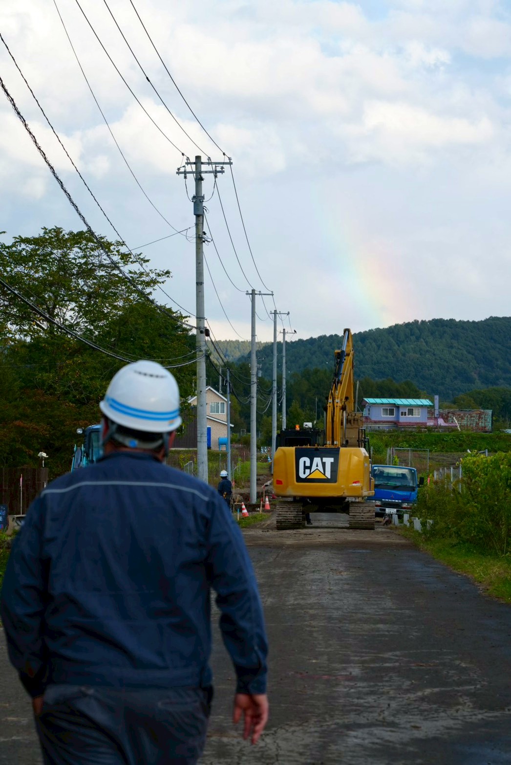 現場へ向かう丸博野沢組のスタッフと建設機械、空には虹